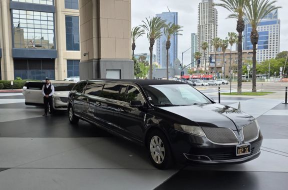 limousine service in vista ca A shiny black stretch limousine from VIP Ride 4 U with a professional chauffeur, parked on a Vista, CA downtown street lined with palm trees and modern high-rise buildings, ideal for executive or event transportation.